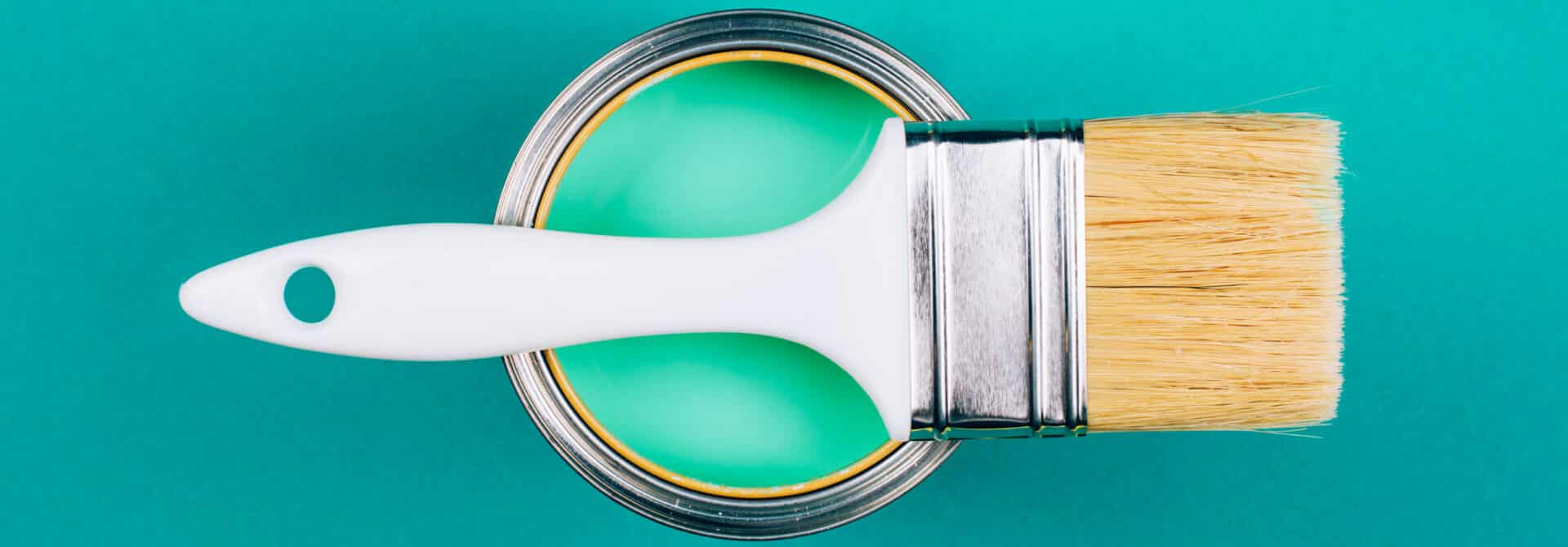 White handle brown bristle paint brush laid horizontally on top of a light blue/green paint bucket with a teal background to represent the types of paint finishes