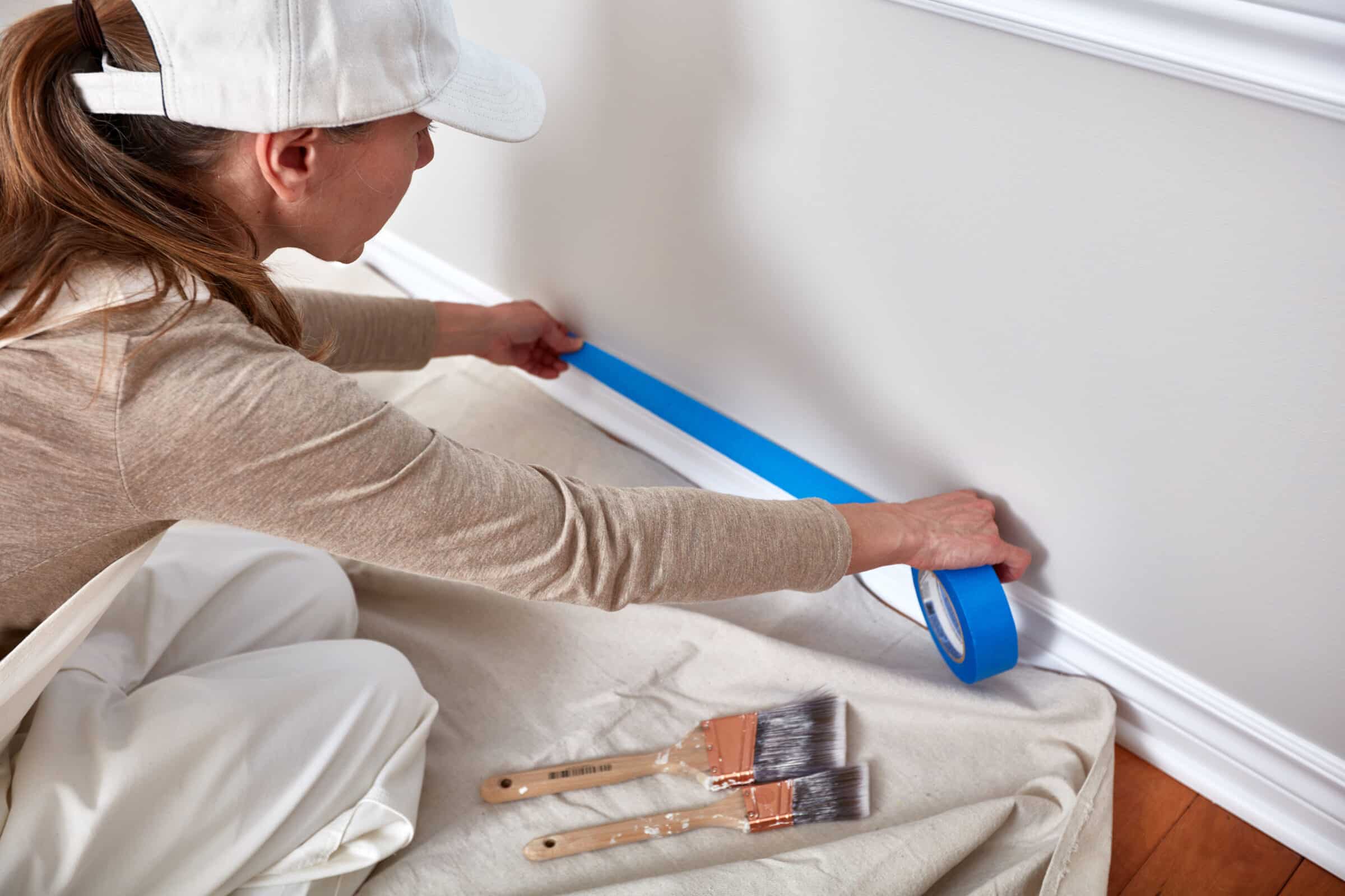 woman with white hat on putting painters tape on her white painted trim near the floor 