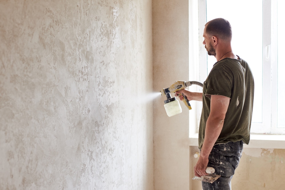 man in paint clothes using a spray gun to paint a interior home's wall with a window behind him