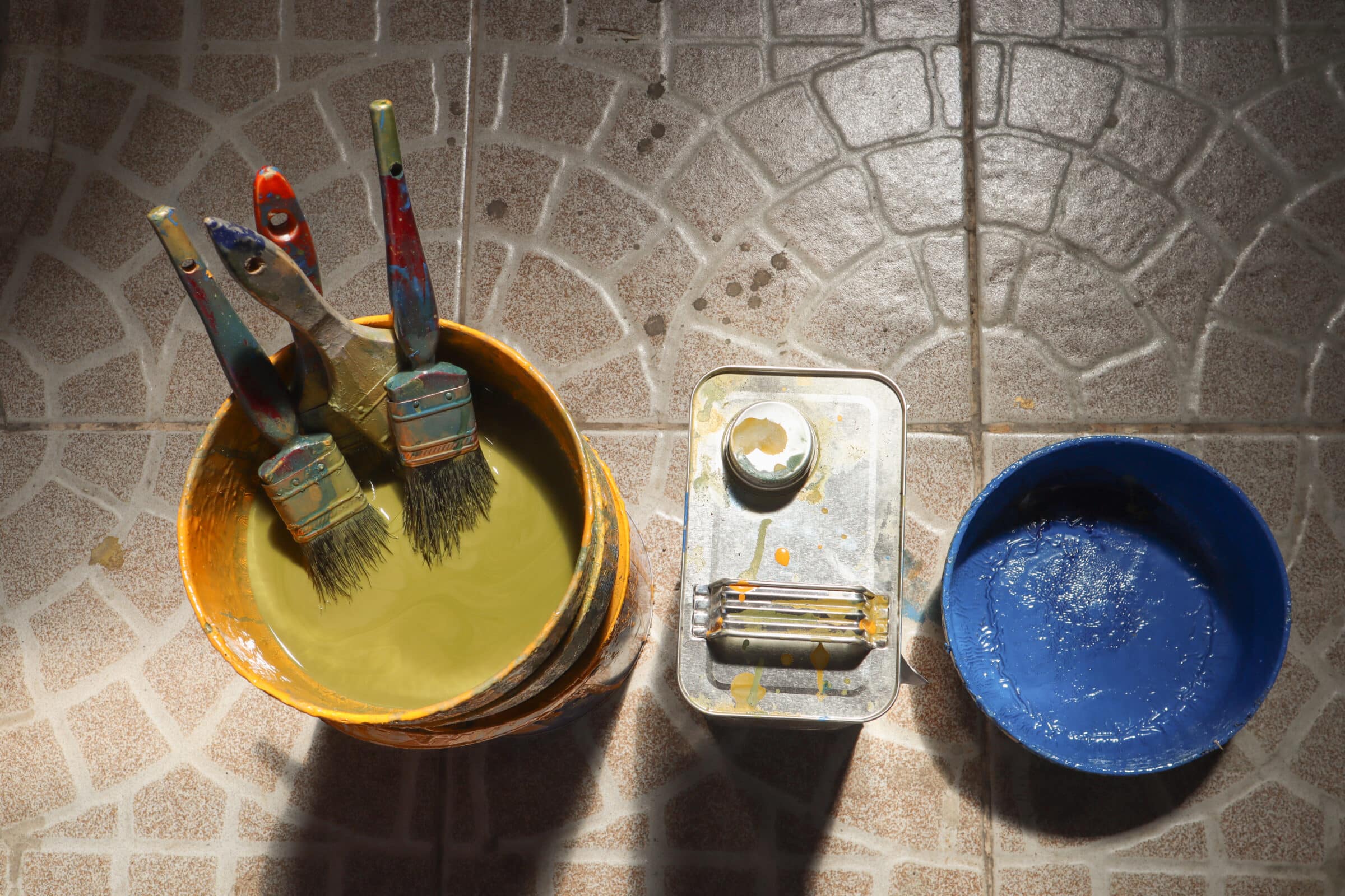 old yellow bucket with 4 dirty paint brushes sitting in a type of paint thinner next to a jug of thinner and a blue bucket of paint on a ceramic beige tile floor 