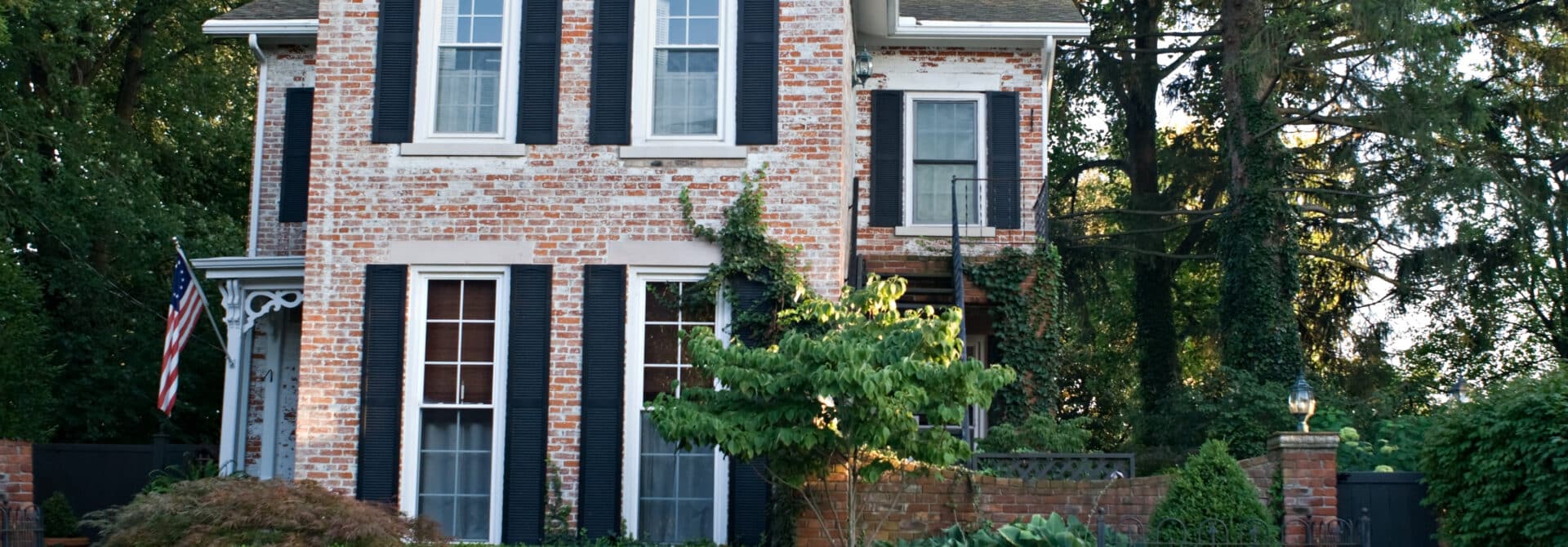 brick house with paint and black shutters with green yard and bushes with a American flag on the porch